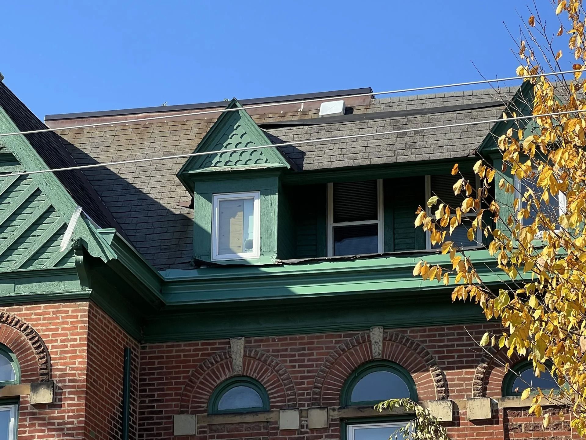 Brick building roof with green dormer windows and autumn tree branches against a blue sky