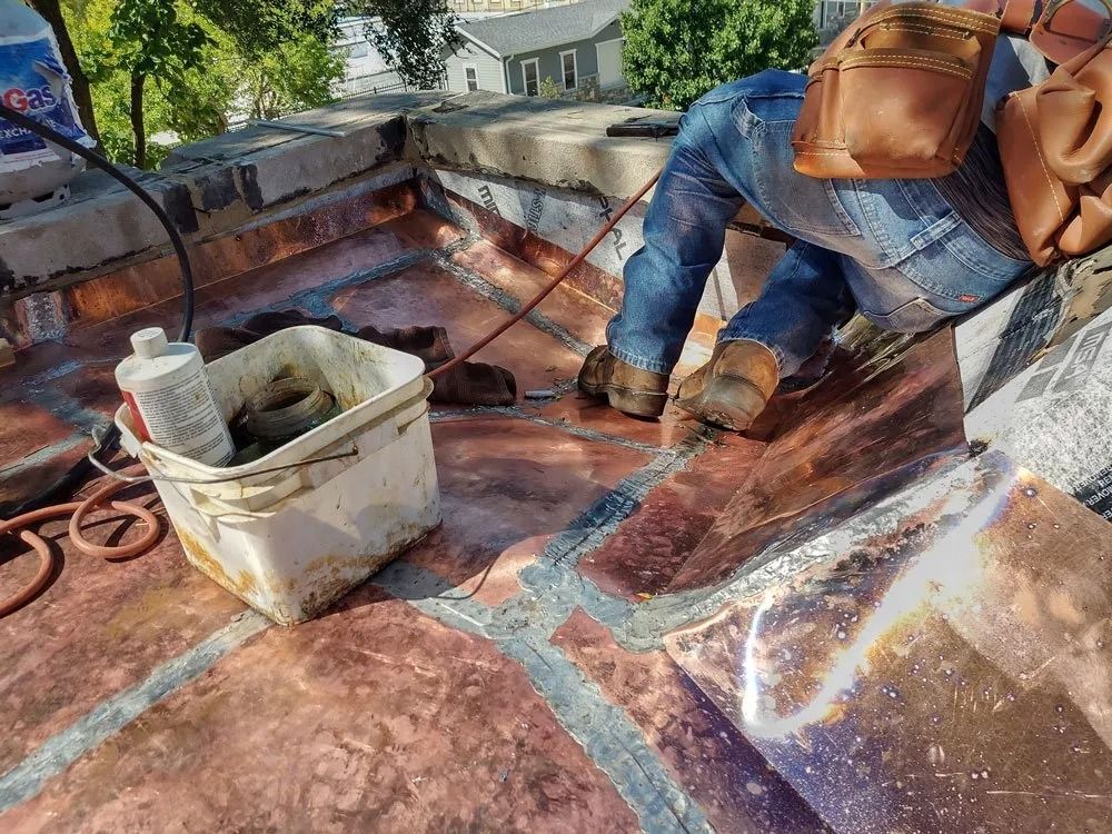 Worker on a rooftop applying sealant beside a bucket and tools near a house.