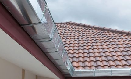 Roof eaves with metal gutters beside a red tiled roof under a cloudy sky