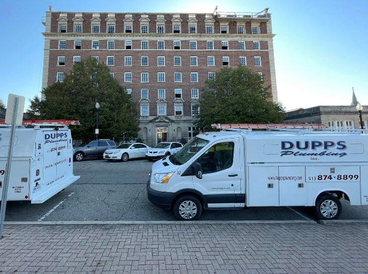 Two white Dupps Plumbing service vans parked in a lot in front of a multistory brick building.