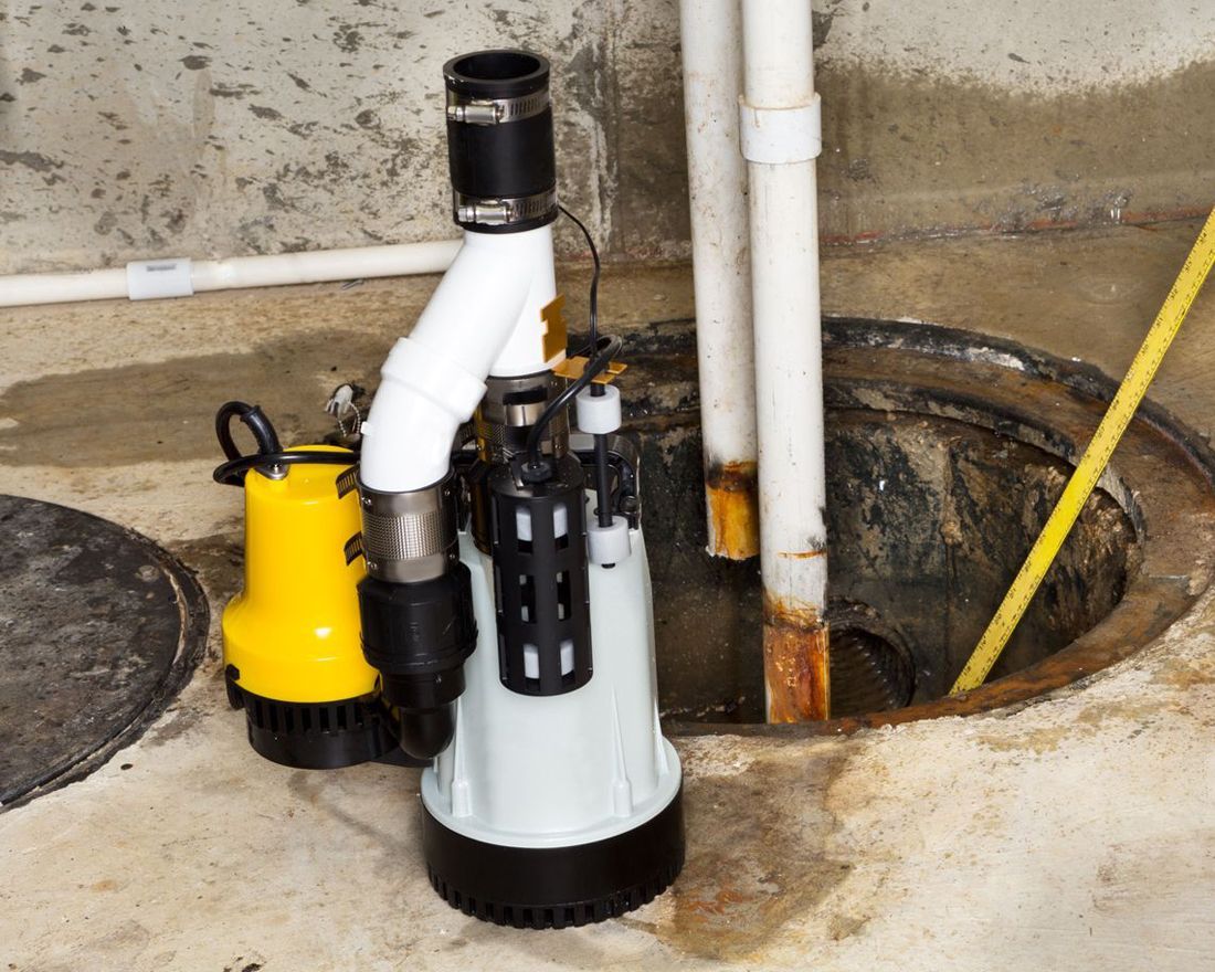 Sump pump and plumbing pipes installed in a basement floor drain pit, with tools nearby.