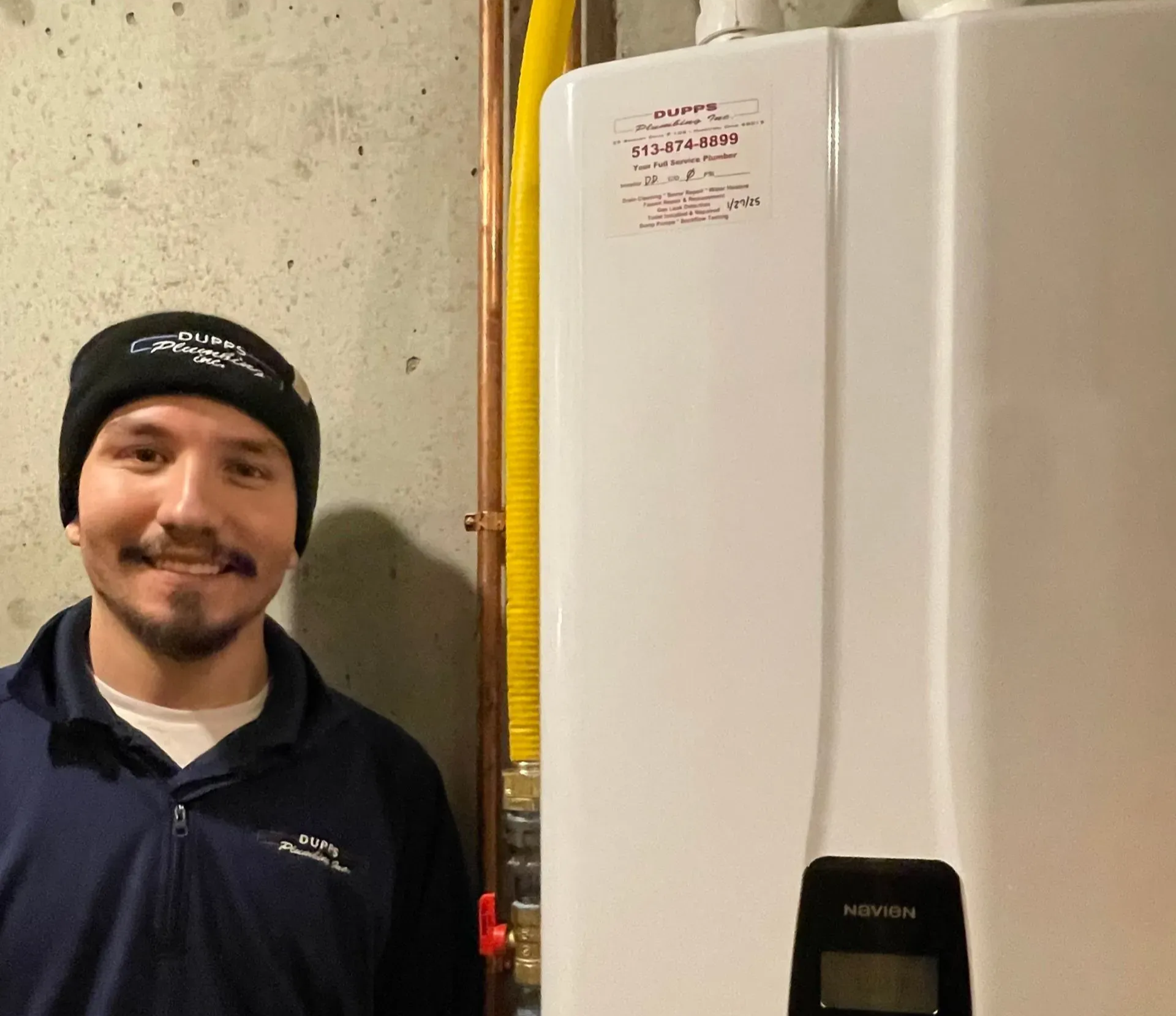 A service professional wearing a branded beanie and jacket stands beside a wall-mounted white tankless water heater.