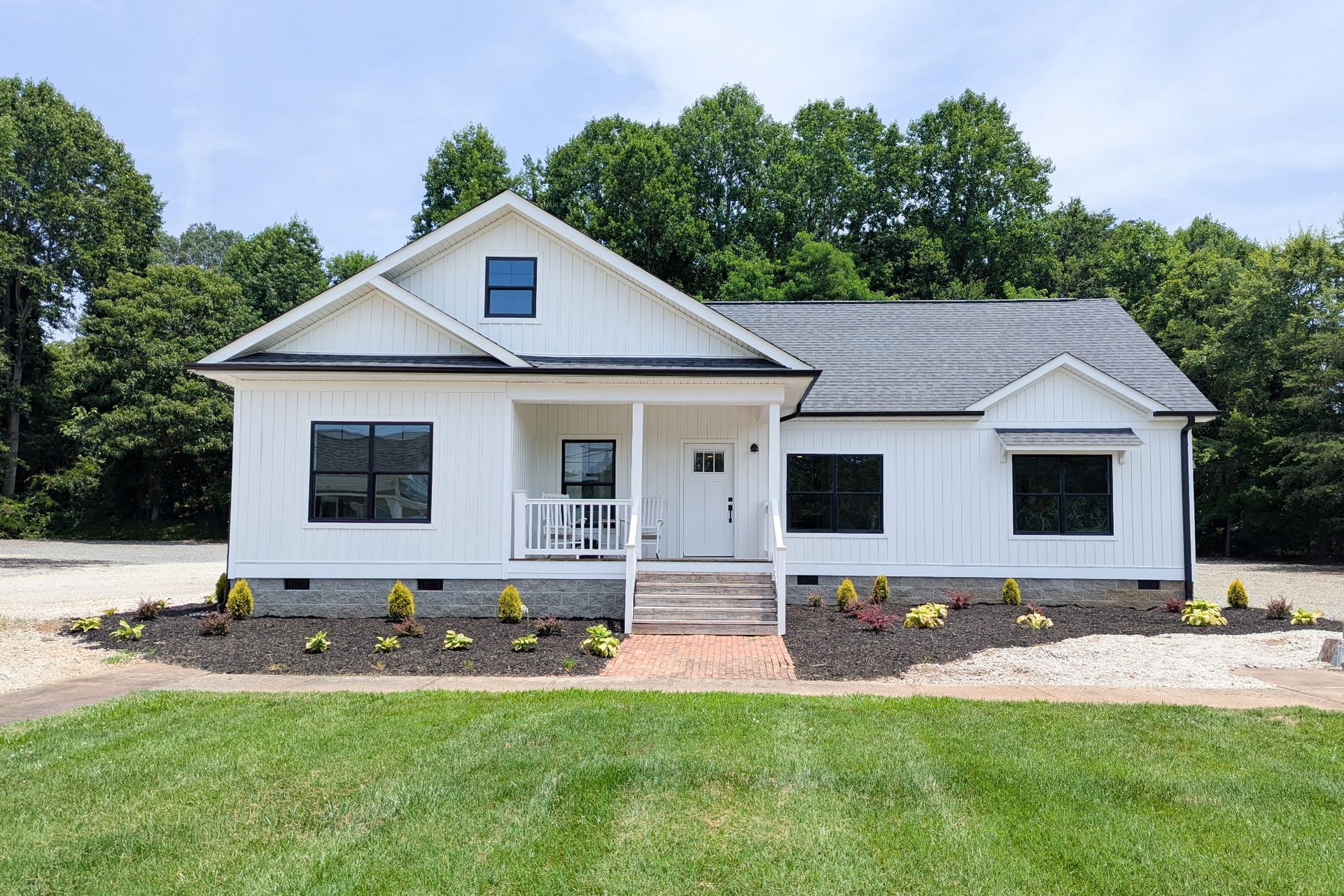 White farmhouse with black windows and a small porch, set against a backdrop of green trees.