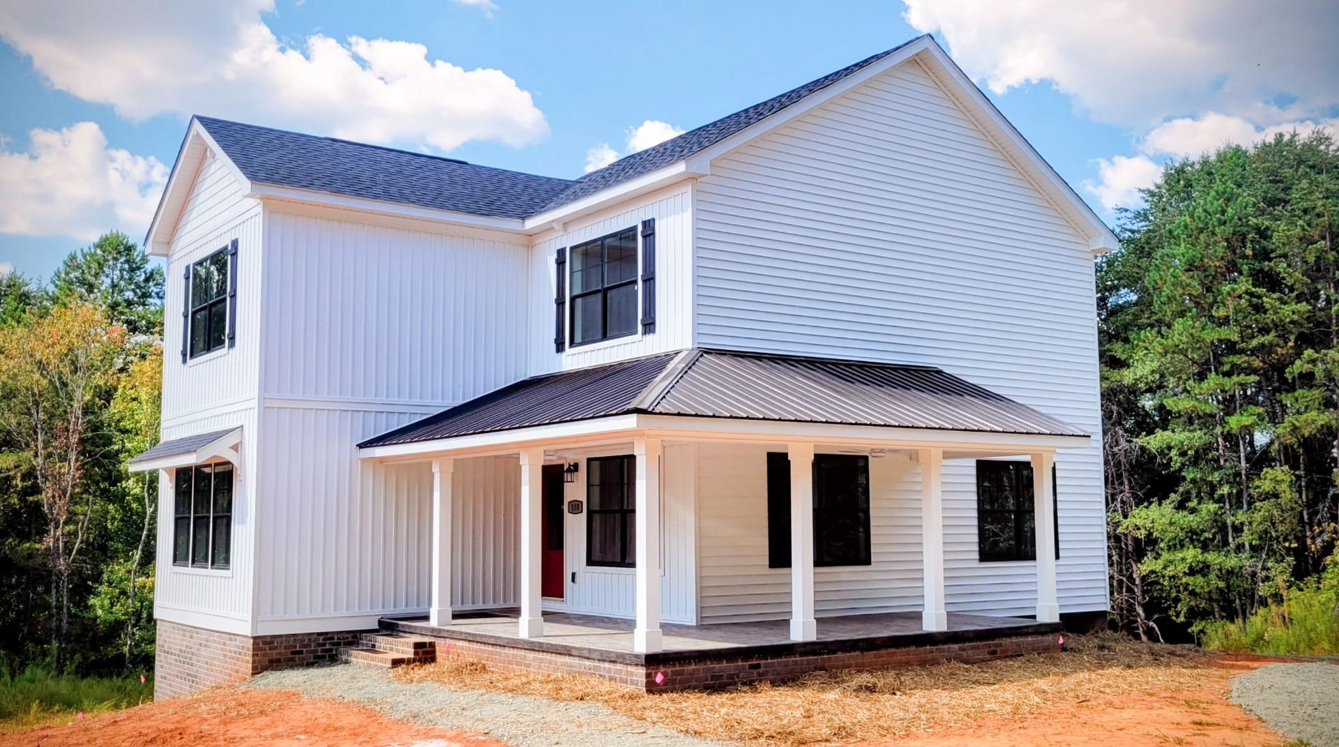 White two-story house with black trim, porch, and metal roof against a sunny sky and trees.