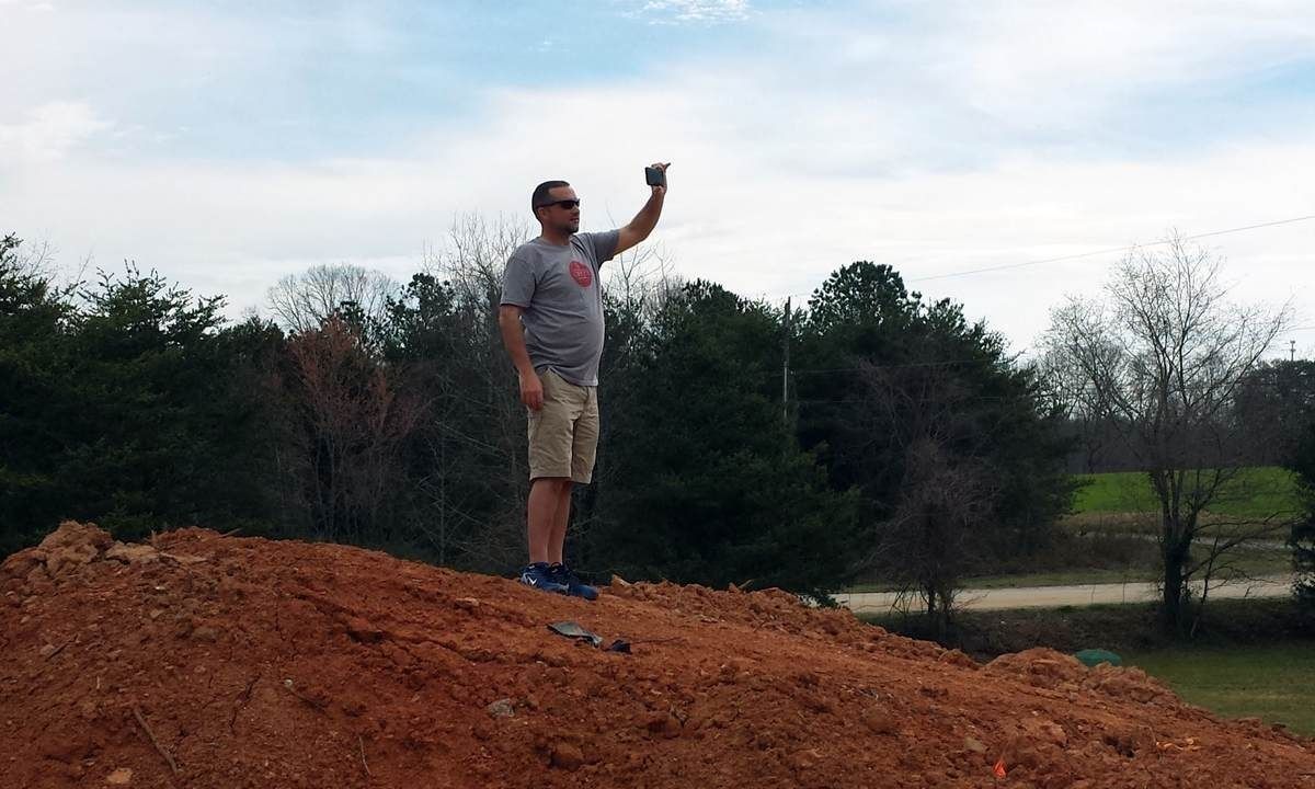Man on a dirt pile holds up a small object; sky, trees, and field in the background.