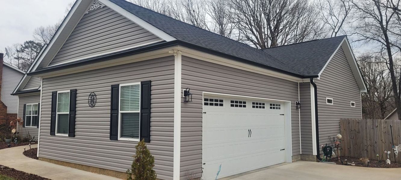 A house with gray siding, black shutters, and a white garage door. Black roof. Cloudy sky.