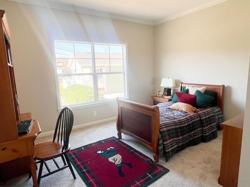 Bedroom with bed, desk, chair, rug, and window, mostly beige and wood tones, some red and green accents.