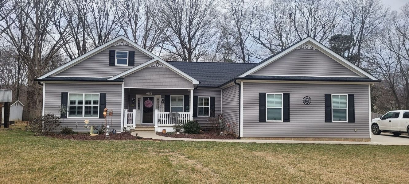 Gray-sided house with black shutters, front porch, and black roof. Bare trees in the background, parked truck on right.