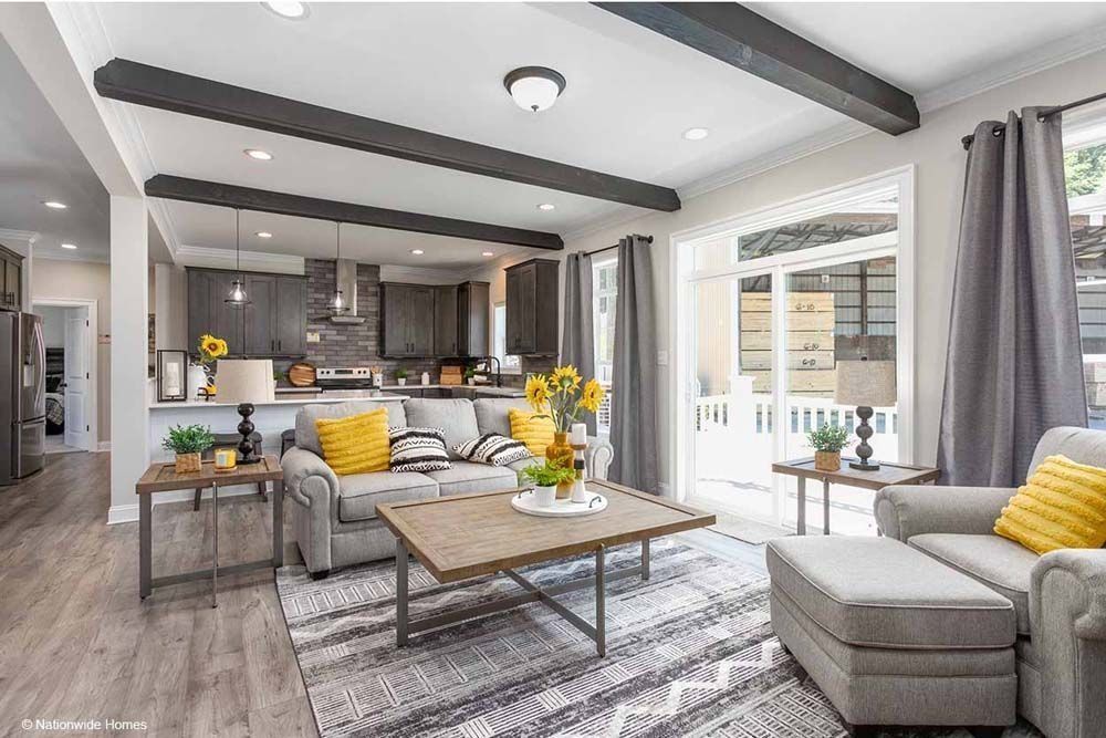 Living room with gray couch, yellow pillows, wooden beams, and kitchen in the background.