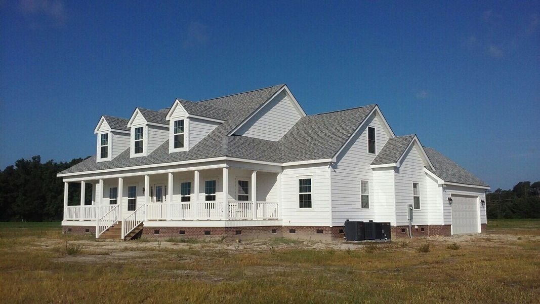 White farmhouse with wrap-around porch, dormers, and gray roof against a clear blue sky.