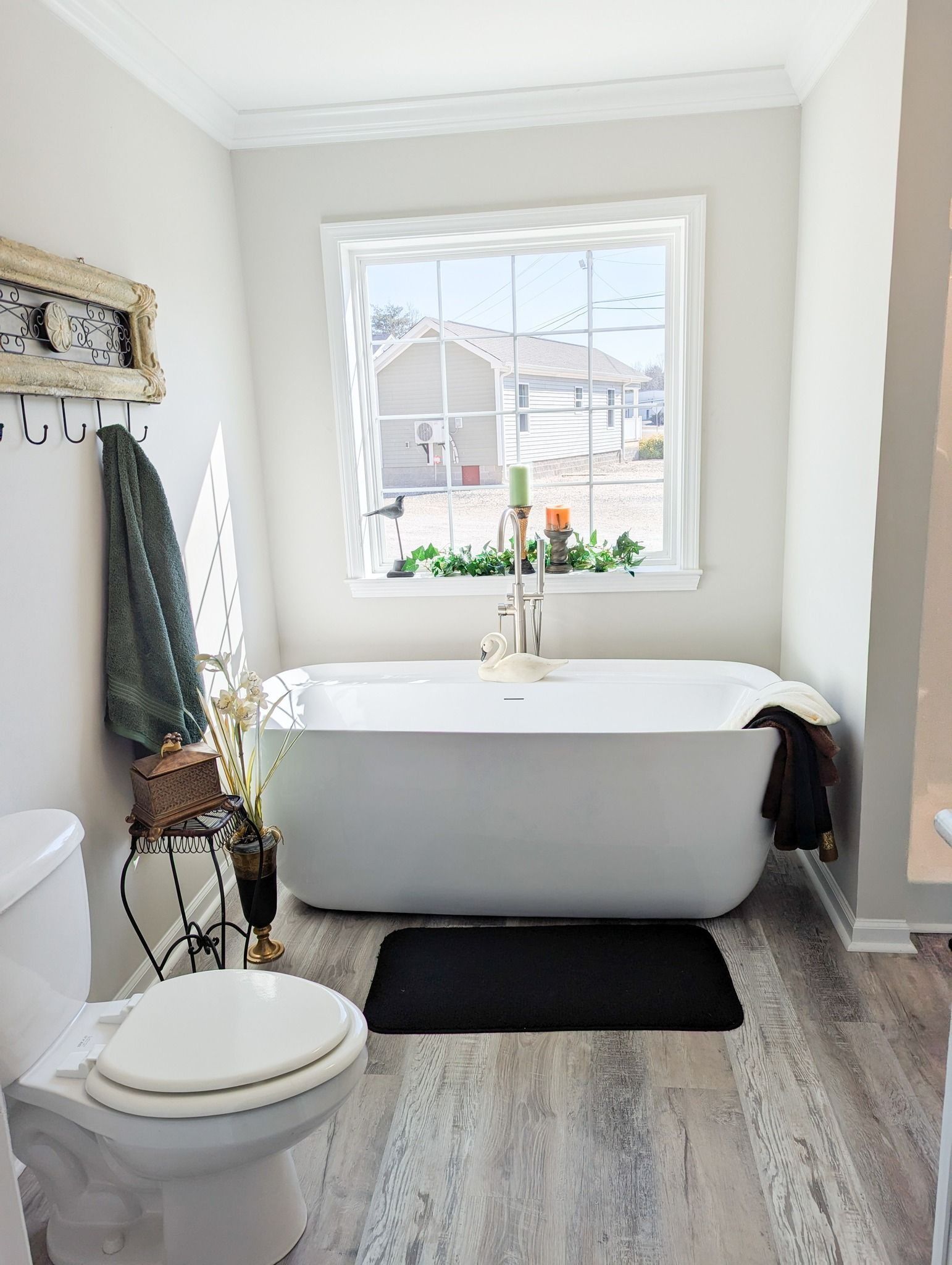 White bathroom with a freestanding tub under a window, toilet, and a black rug.