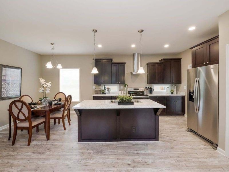 Spacious kitchen with island, dining table, dark cabinets, stainless steel fridge, and light-colored wood-look flooring.