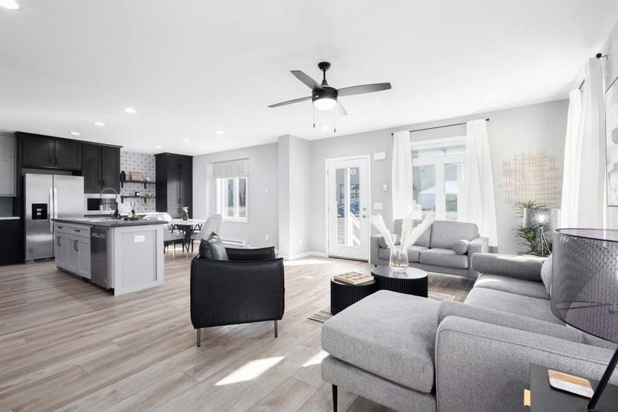 Open-concept living space. Kitchen with dark cabinets, gray island; gray sofa, black chairs, light wood flooring.