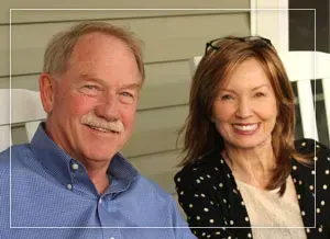 Smiling older couple sitting on a porch. Man in blue shirt, woman in polka dots.