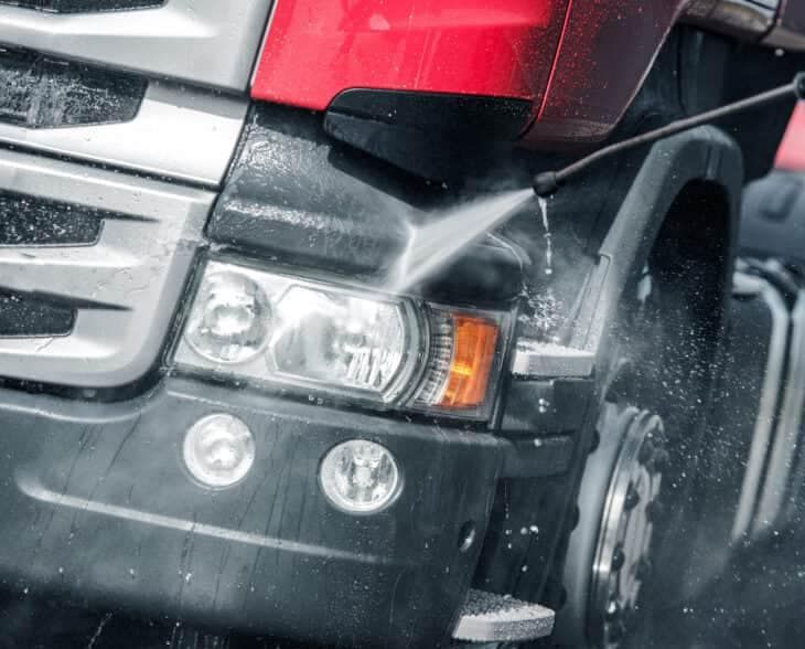 Pressure washing a red truck's front end, spraying water on the bumper, headlights, and tire.
