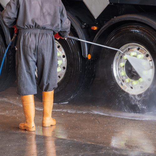 Person in coveralls washing a truck tire with high-pressure water in a concrete setting.
