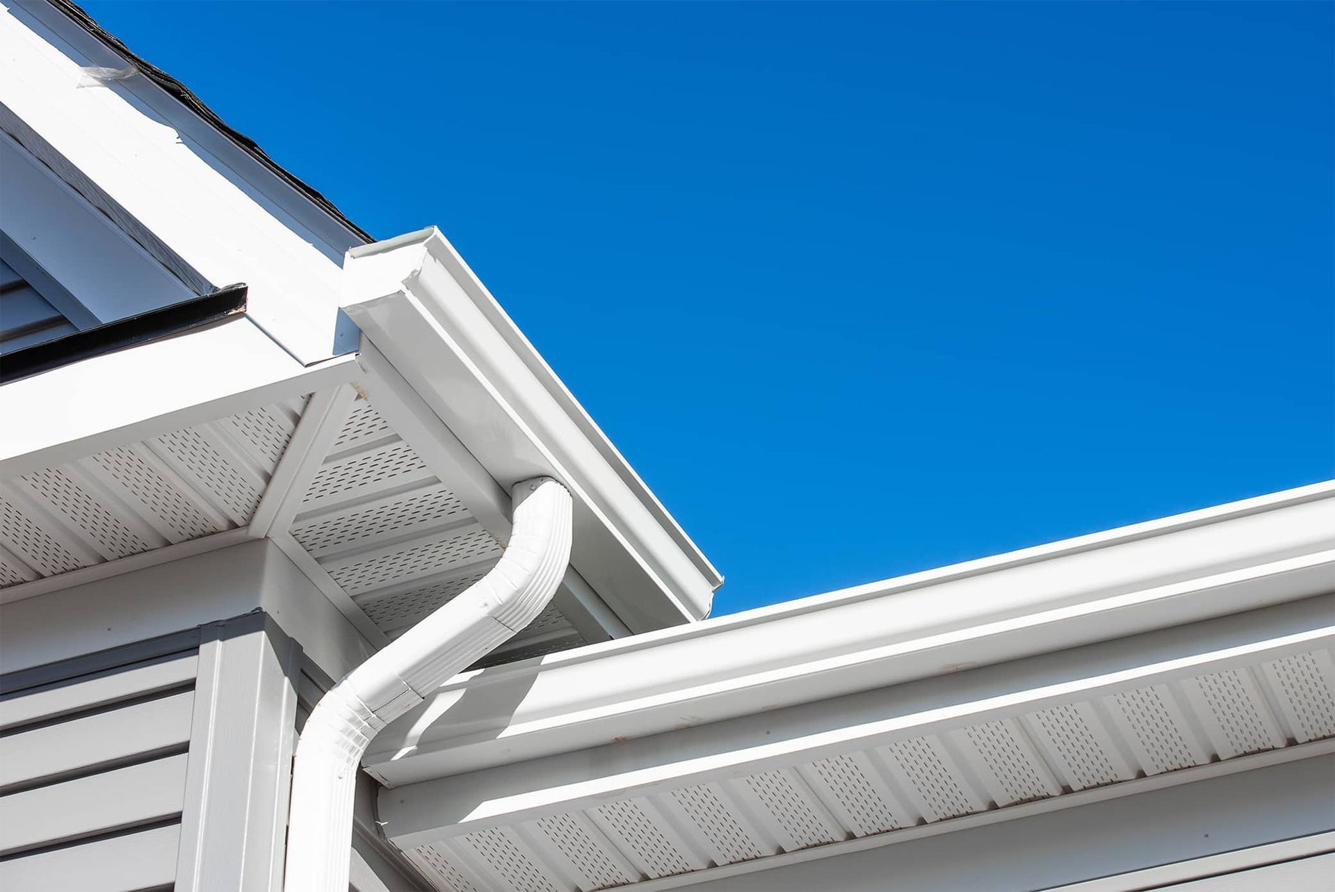 White gutters and downspout on a house with gray siding against a bright blue sky.