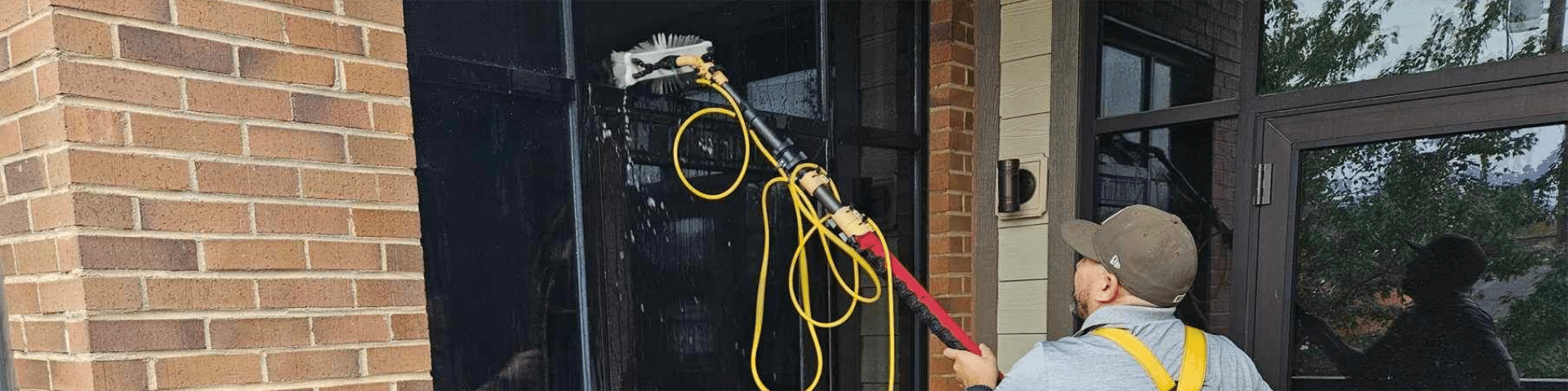 A person using a water-fed pole to wash a black door on a brick building.
