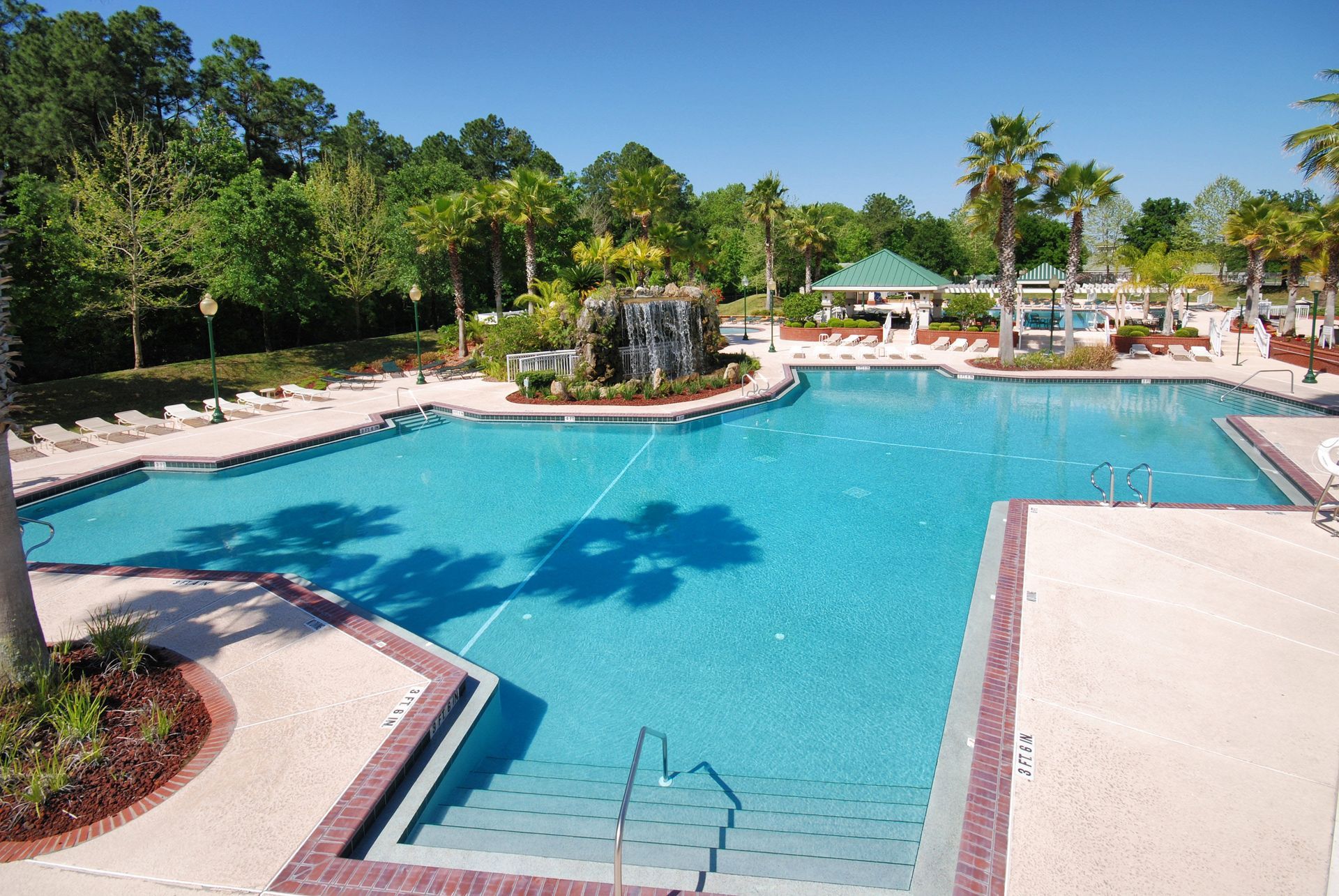 Large swimming pool surrounded by palm trees, greenery, and a building under a blue sky.