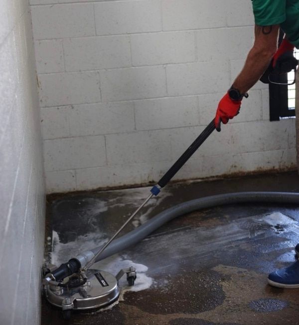 Person using a surface cleaner on a concrete floor inside a white-walled room, with soap and a hose visible.
