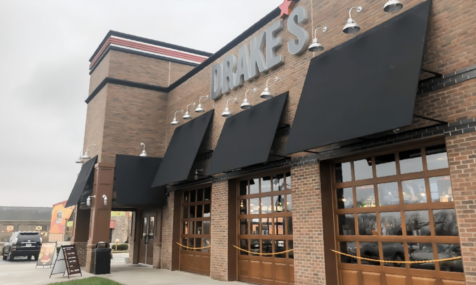 Drake's restaurant exterior with brick walls, black awnings, and garage-style doors. Cloudy sky.