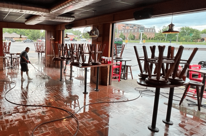 A person power washes a wet, wooden-floored outdoor bar area. Tables have chairs stacked on top.