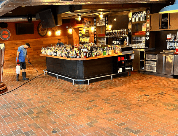 Person spraying the outdoor patio floor of a bar. Bar with bottles visible, brick floor, dark interior.