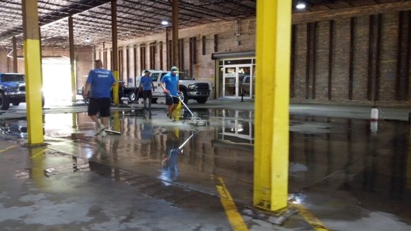 Men mop a flooded industrial space with yellow support beams and vehicles.