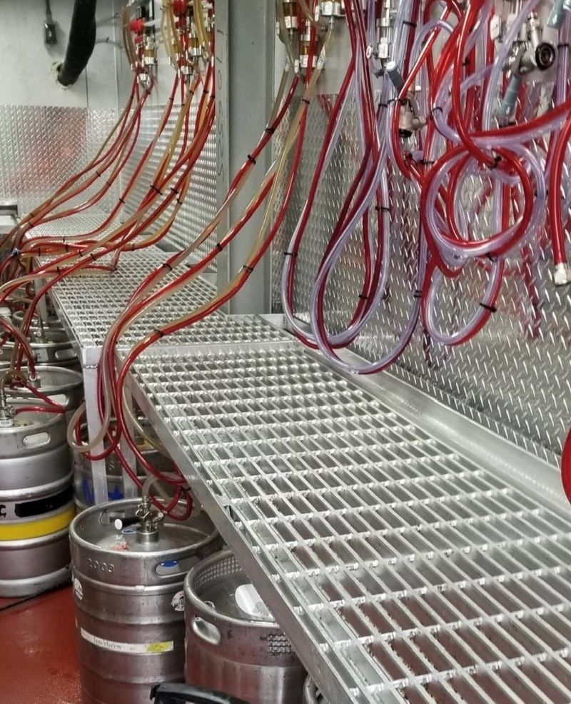 Beer kegs beneath a tap system with red tubing and a metal grated floor.