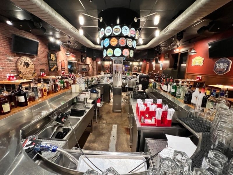 Interior view of a bar with a long counter, liquor bottles, and tap handles overhead. Red brick walls and TVs.