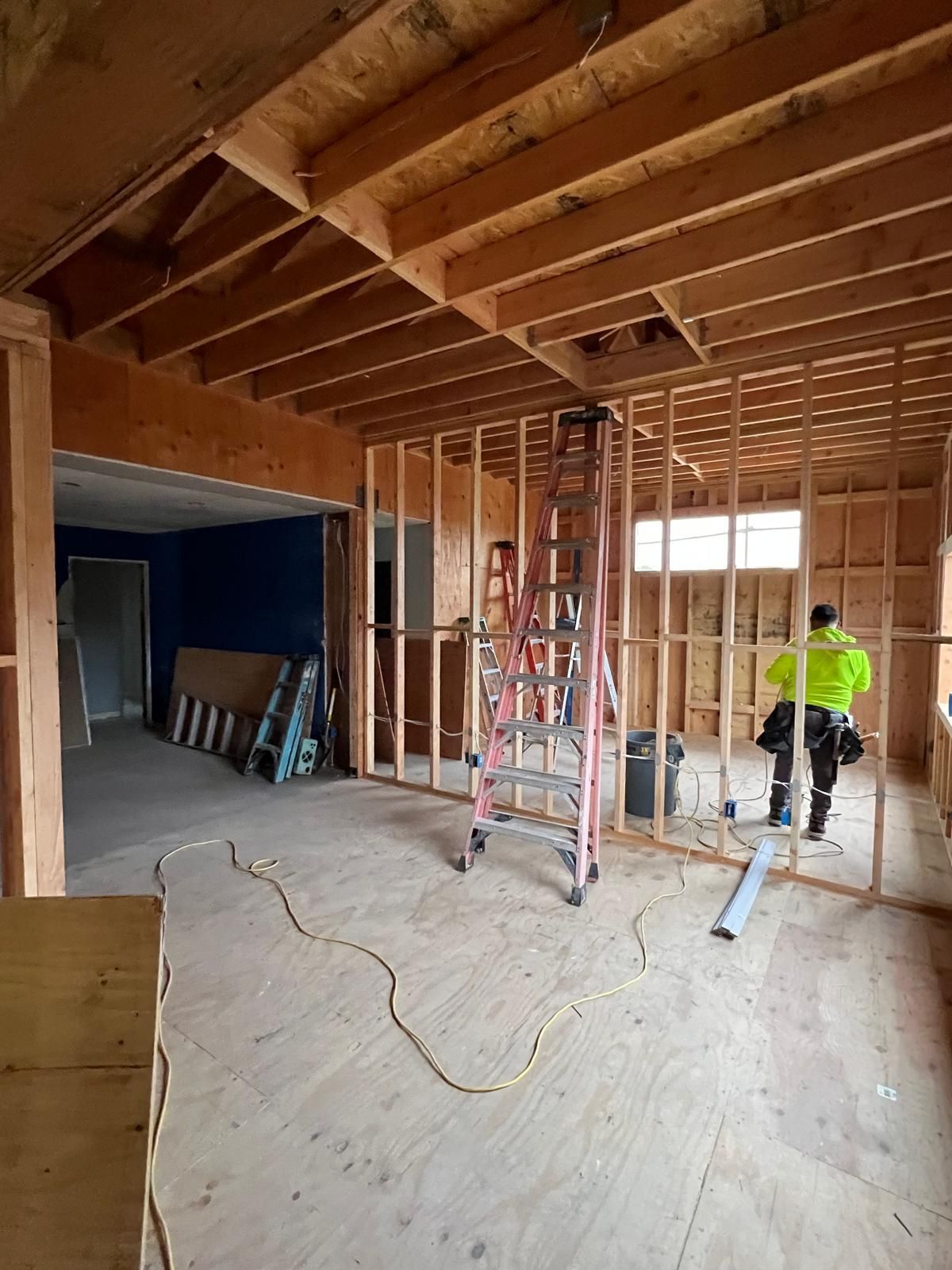 Interior house framing during construction worker in neon vest, ladder, plywood flooring.