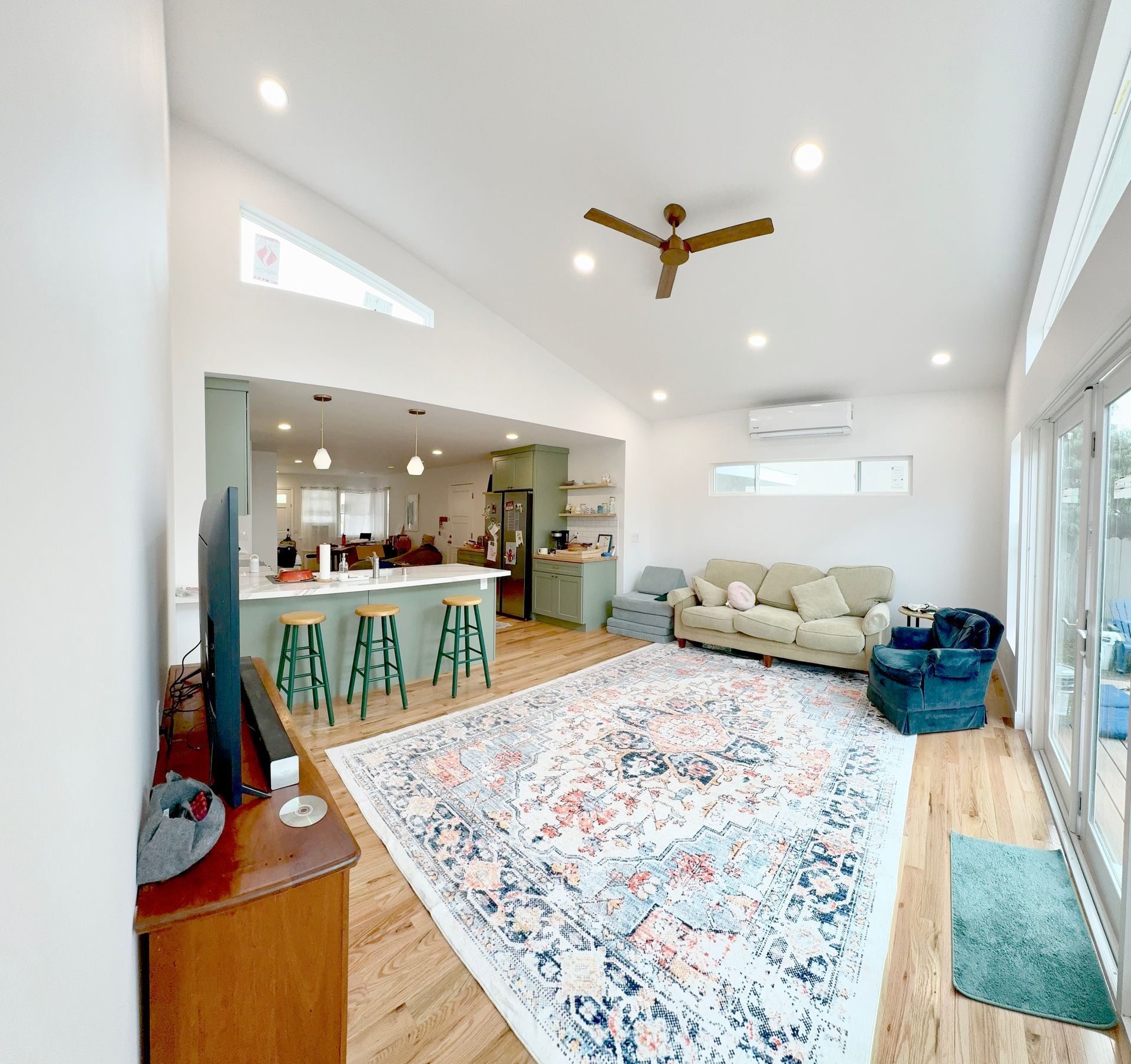 Spacious, bright living area with kitchen. Features hardwood floors, large rug, green stools, and a ceiling fan.