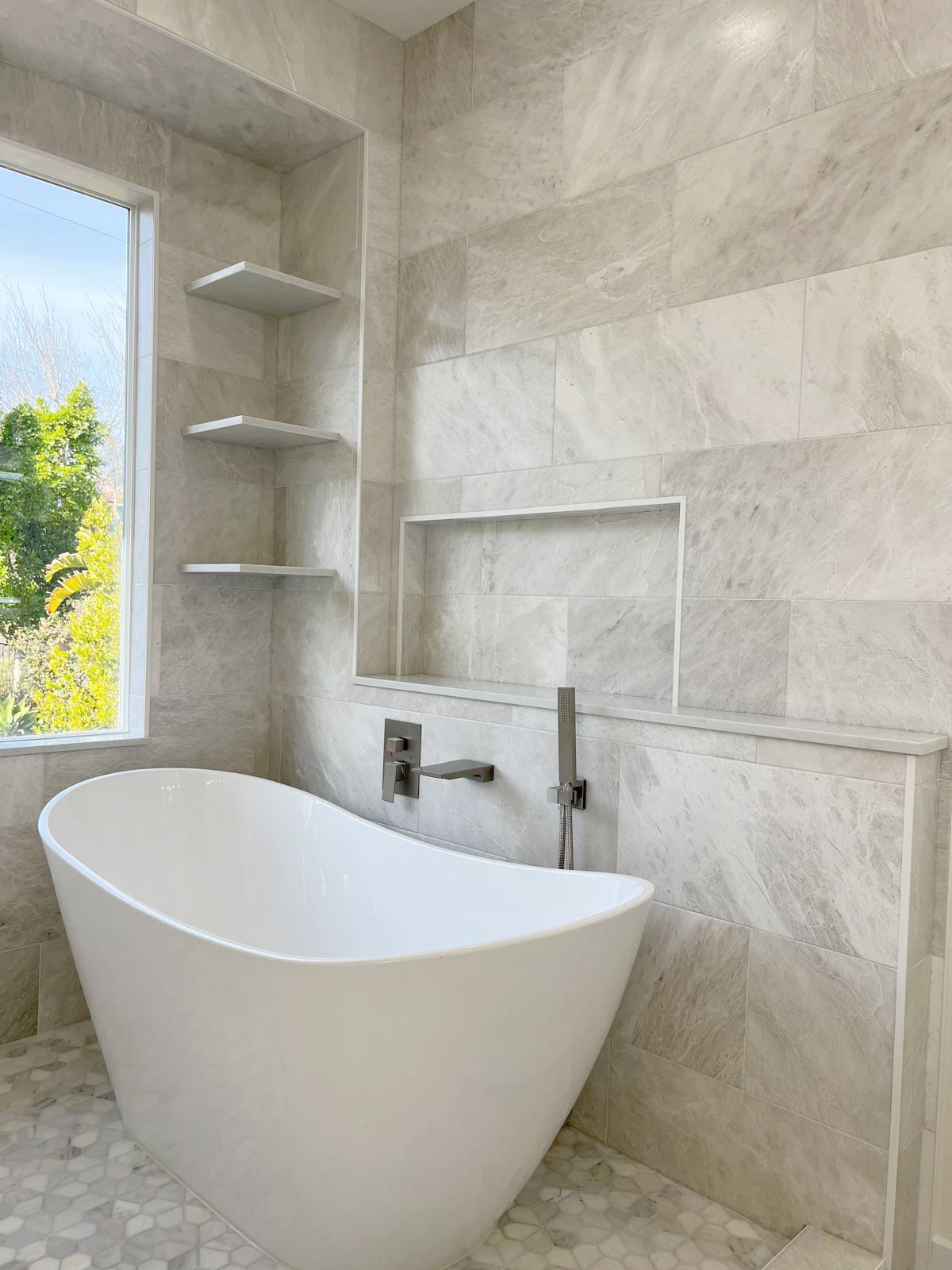 Freestanding white bathtub in modern bathroom with light gray tiled walls and shelves.