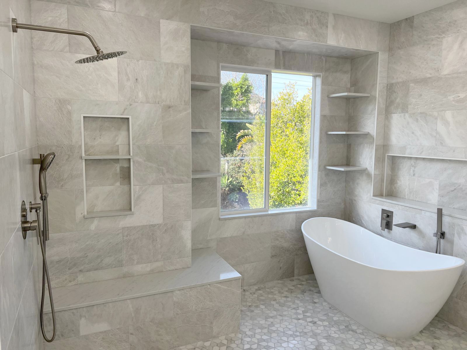 Modern bathroom with a walk-in shower, freestanding tub, window, and built-in shelves. Gray tiles dominate.