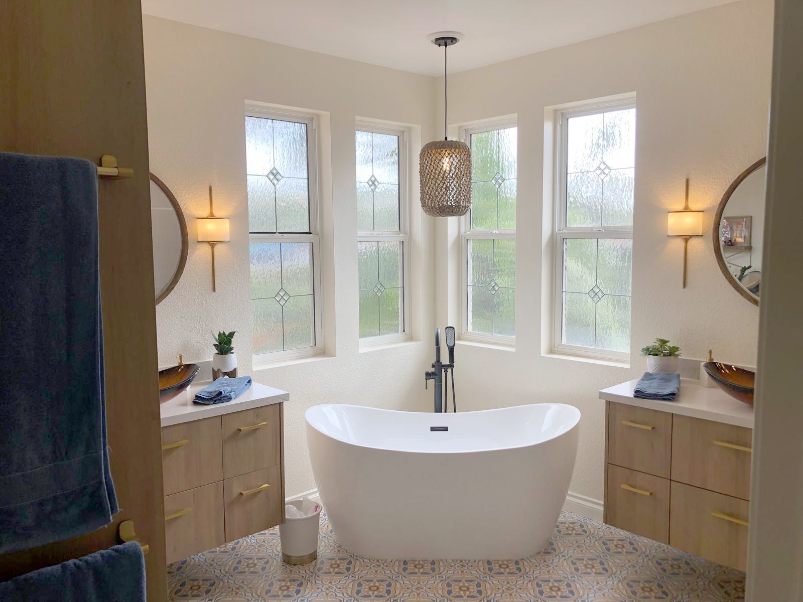 Bathroom with freestanding tub, two vanities, and decorative windows. Light wood cabinets, gold fixtures.