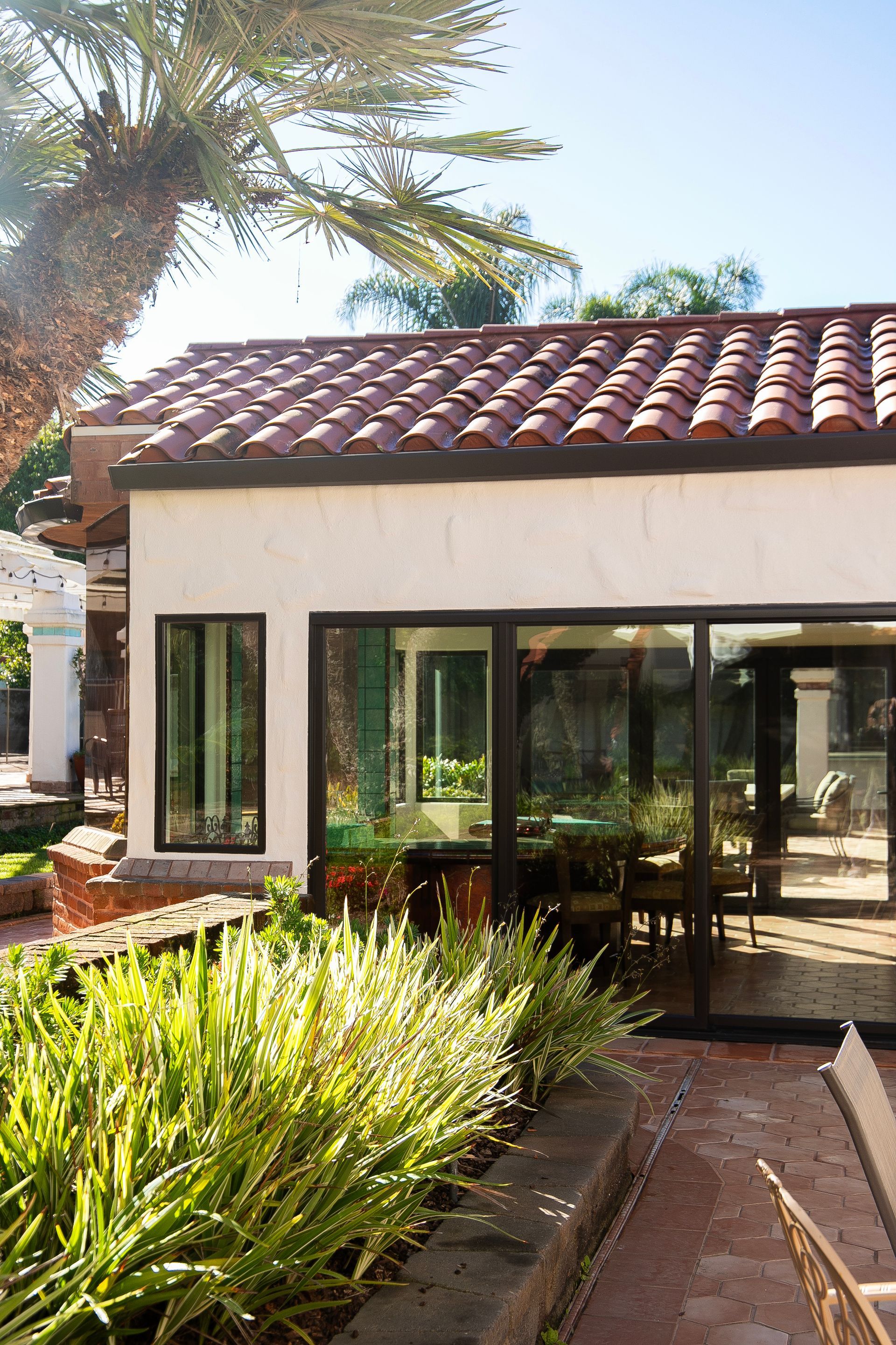 Exterior view of a building with a red tile roof, white stucco walls, and large windows. Palm tree in background.