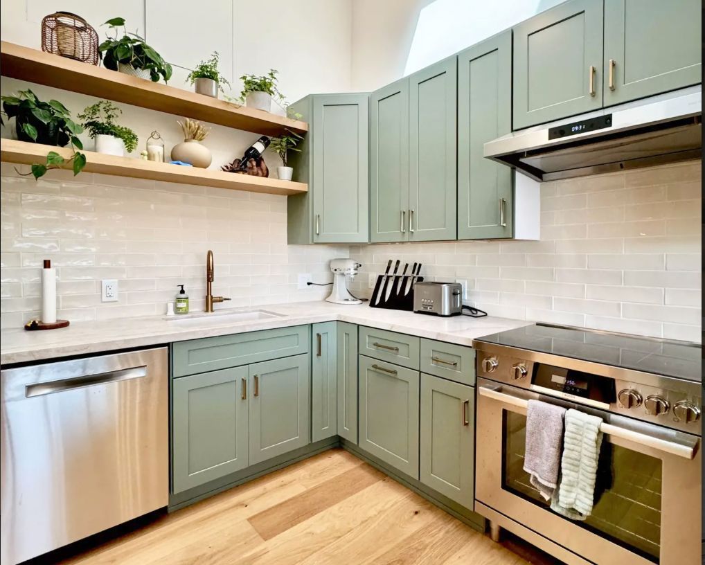 Sage green kitchen with floating shelves, plants, stainless steel appliances, and light wood floors.