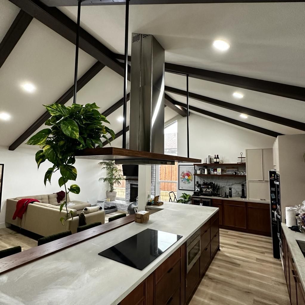 Modern kitchen with a large island, stainless steel hood, wooden beams, and a hanging plant.
