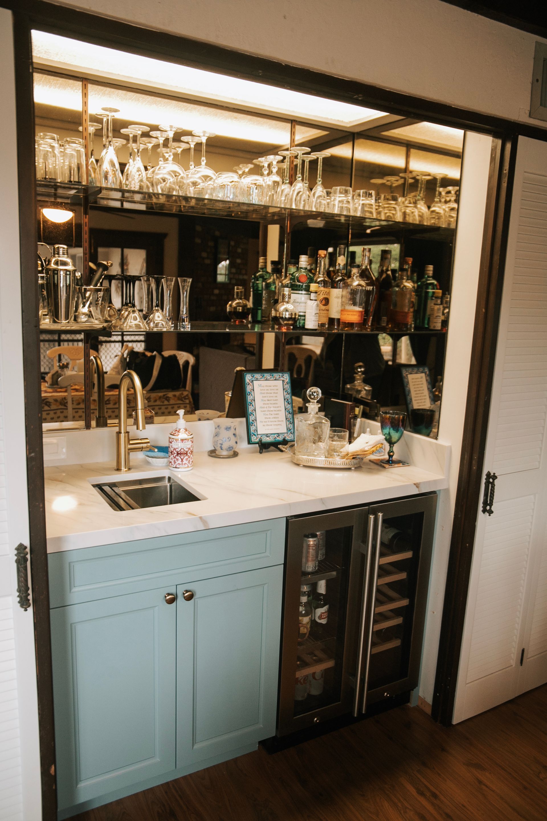 Home bar with sink, wine fridge, glassware, liquor bottles, and mirrored back wall.