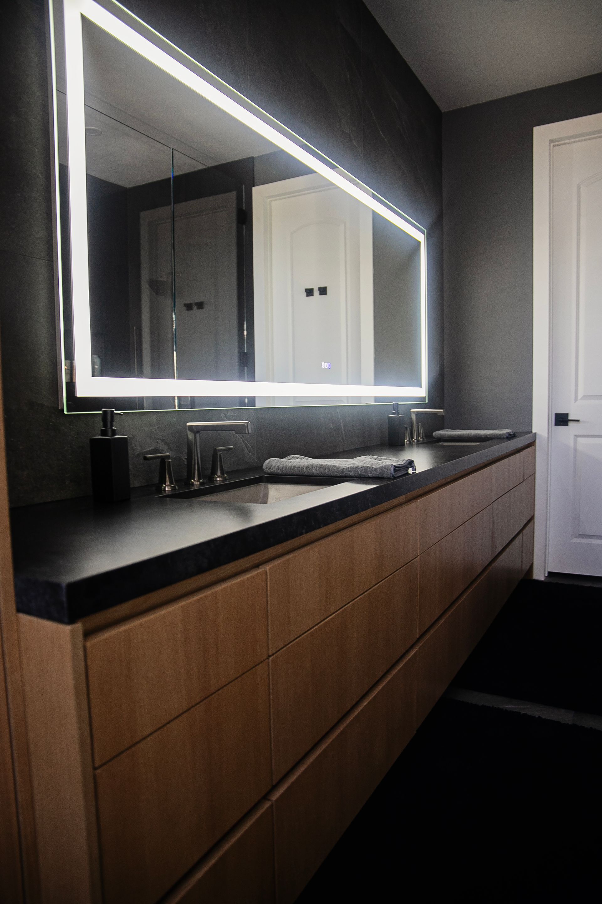 Modern bathroom with a large, lit mirror above a long wooden vanity with a black countertop.