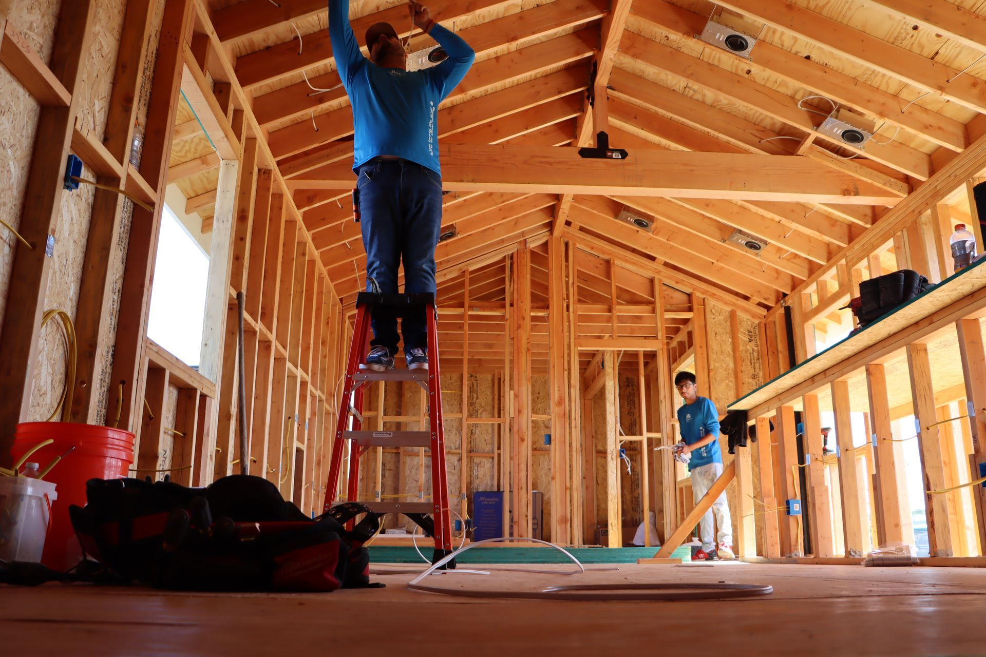 Two workers installing electrical wiring inside a wood-framed building under construction.