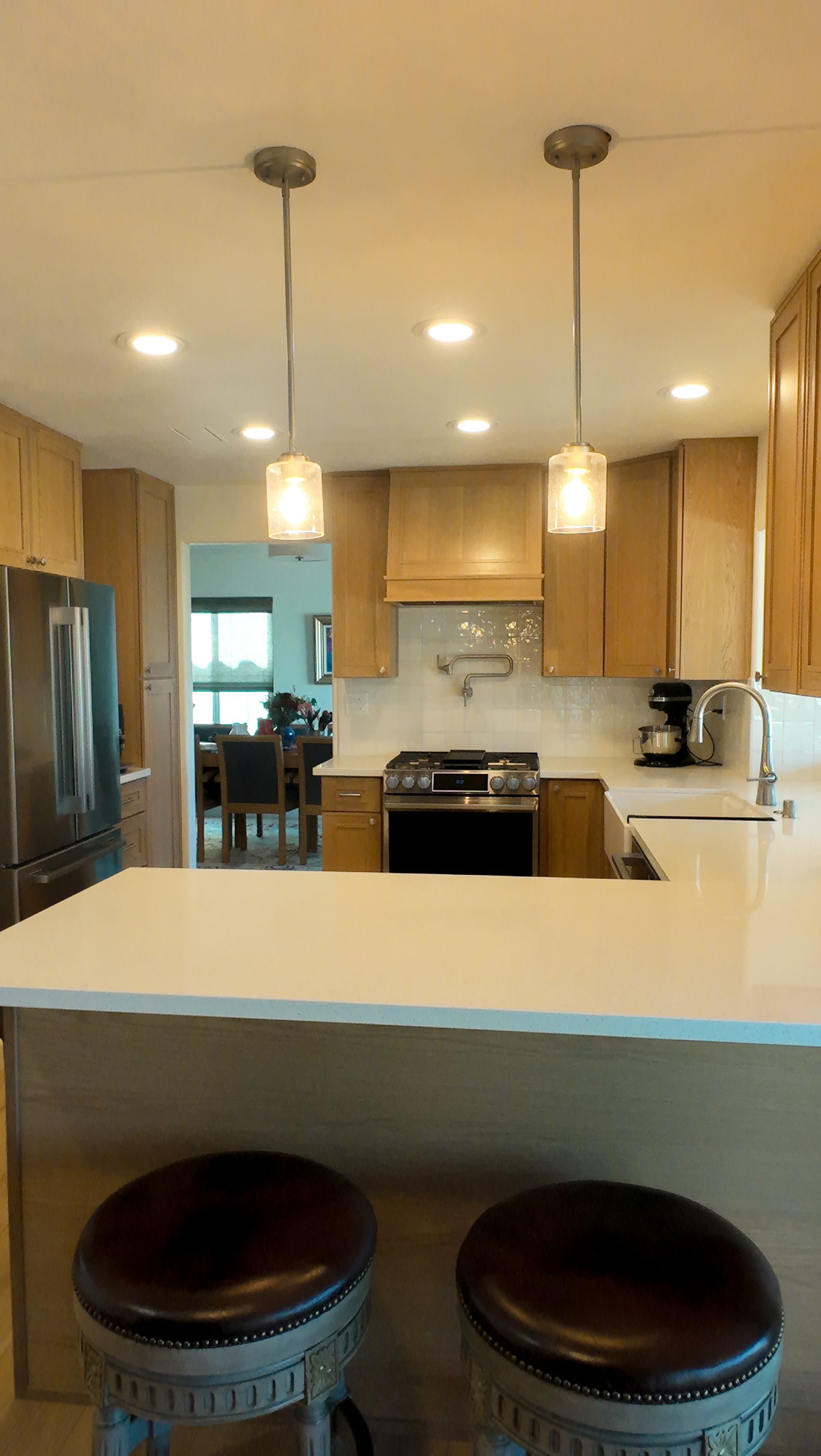 Kitchen with two pendant lights, light wood cabinets, and a white countertop. Two bar stools are in the foreground.