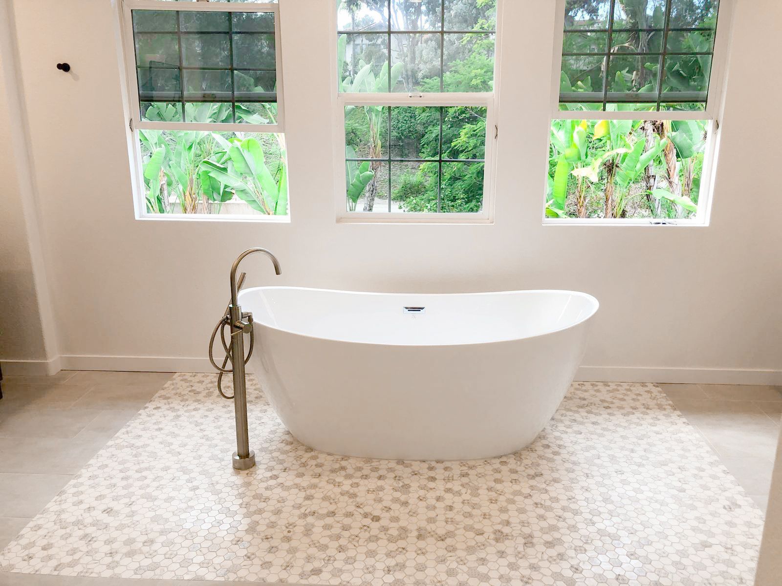 White bathtub in a bright bathroom with three windows overlooking lush greenery.