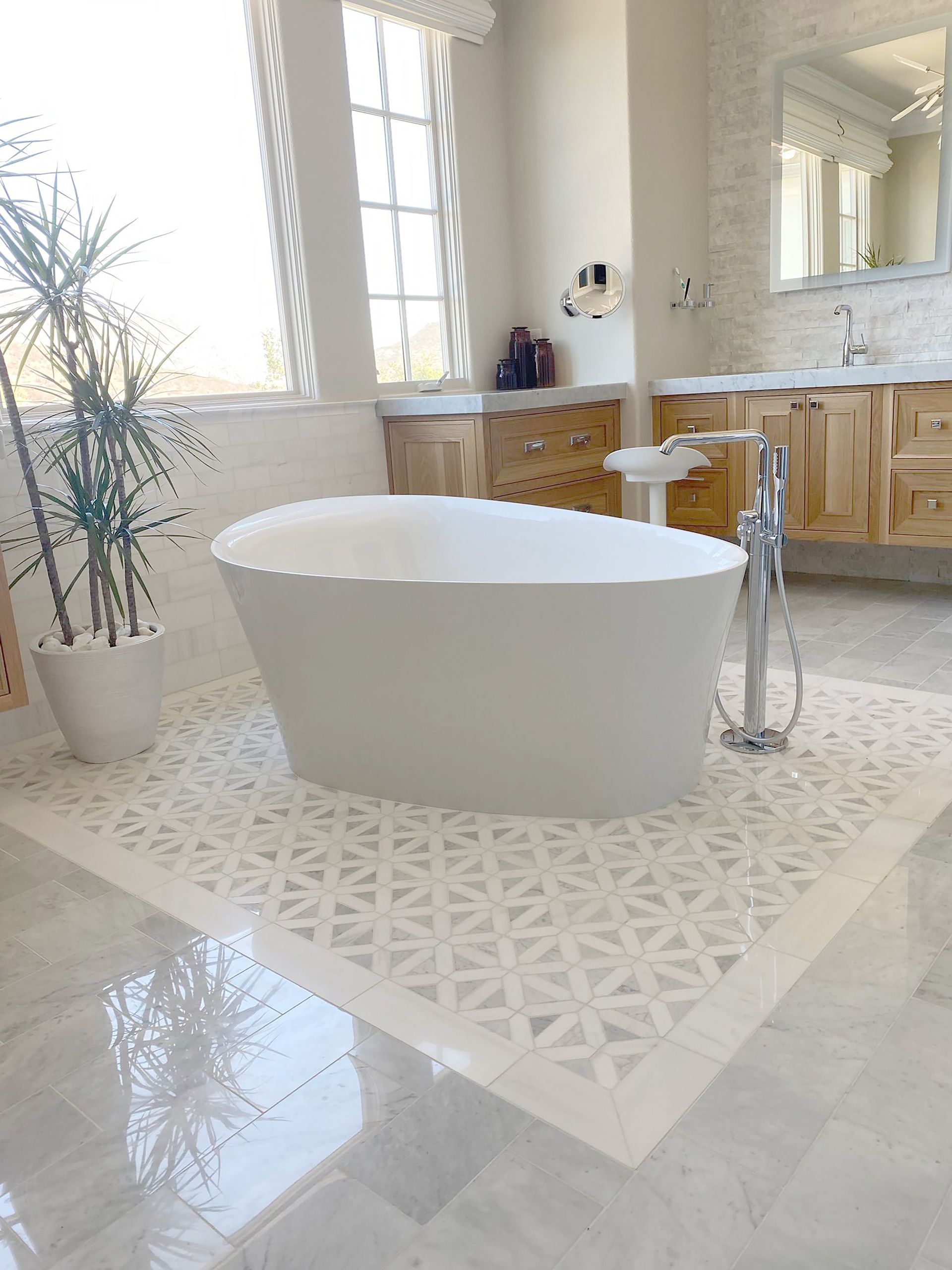 White bathtub in a spacious bathroom with patterned tile flooring, wood cabinets, and large window.