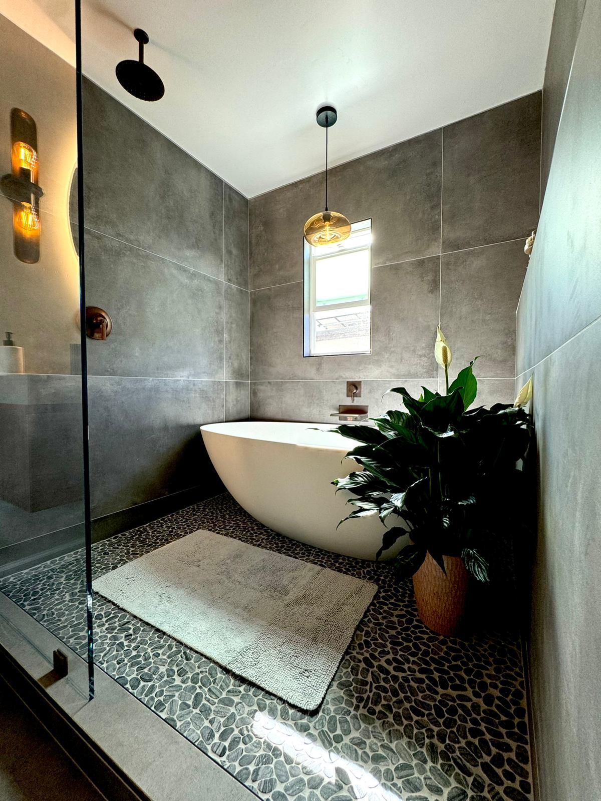 Bathroom with gray tile walls, a white tub, and pebble floor. A large plant sits near the tub.