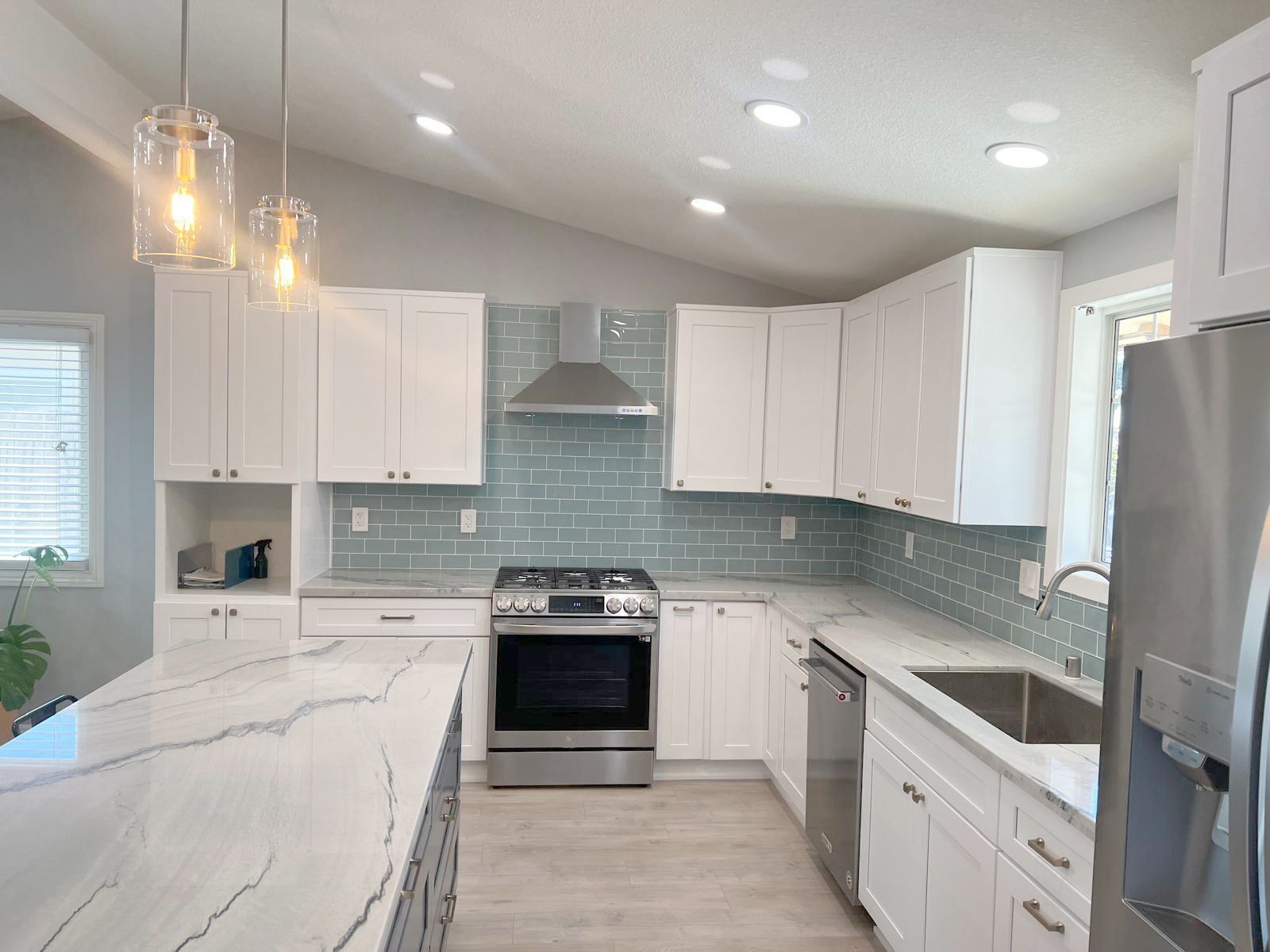 Modern white kitchen with island, blue tile backsplash, stainless steel appliances, and pendant lights.
