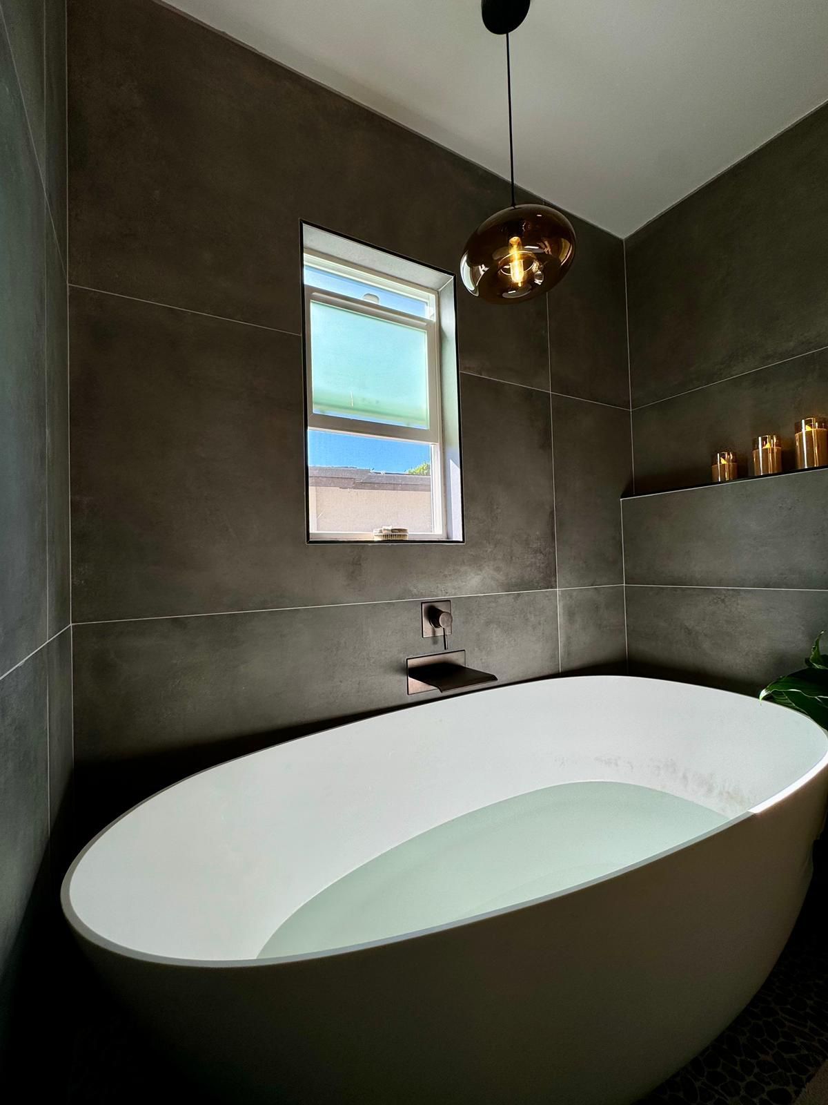 Modern bathroom with oval bathtub filled with water; gray walls, window, pendant light.