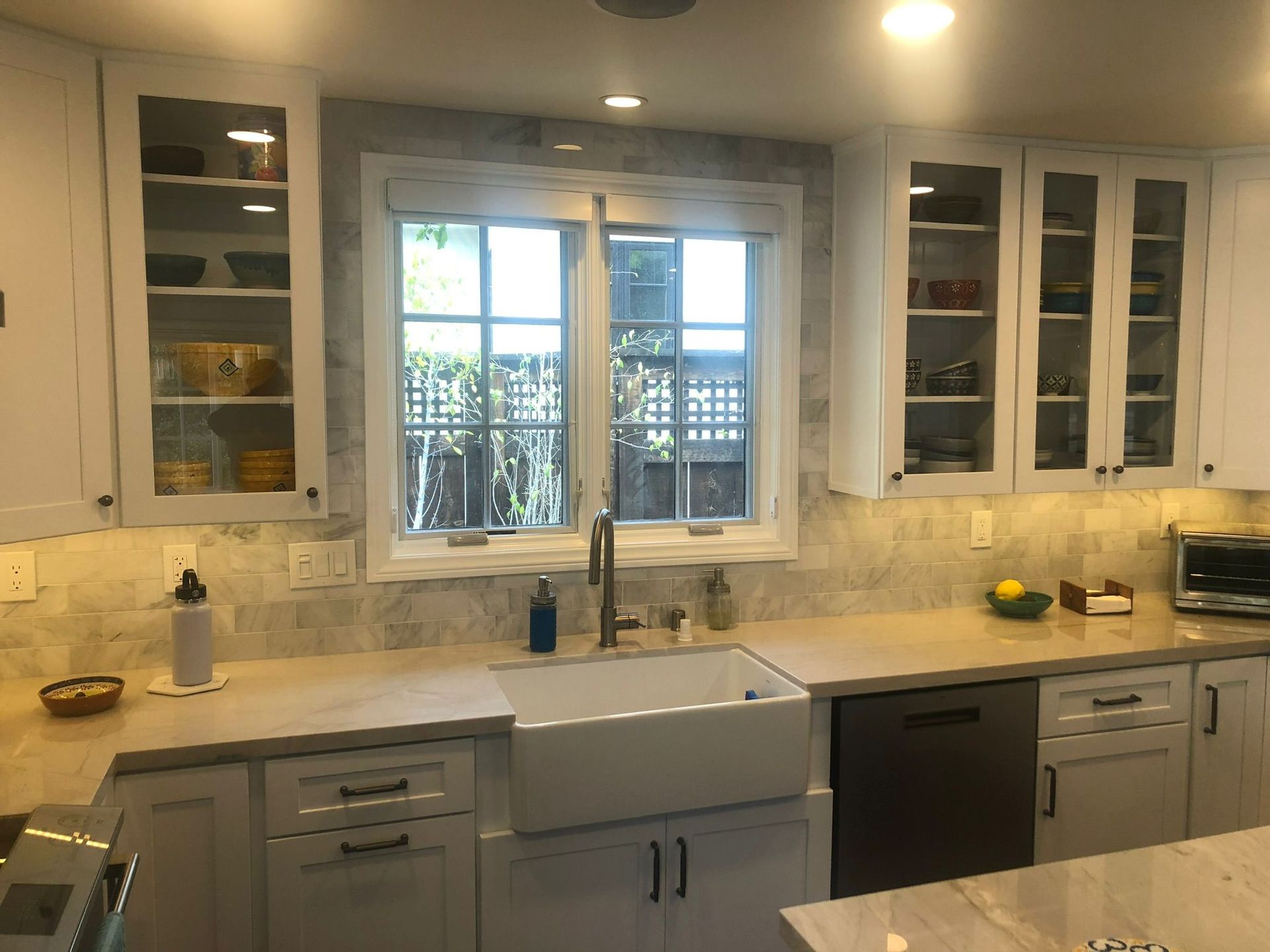 White kitchen with marble backsplash, cabinets, and a farmhouse sink. Window over the sink.