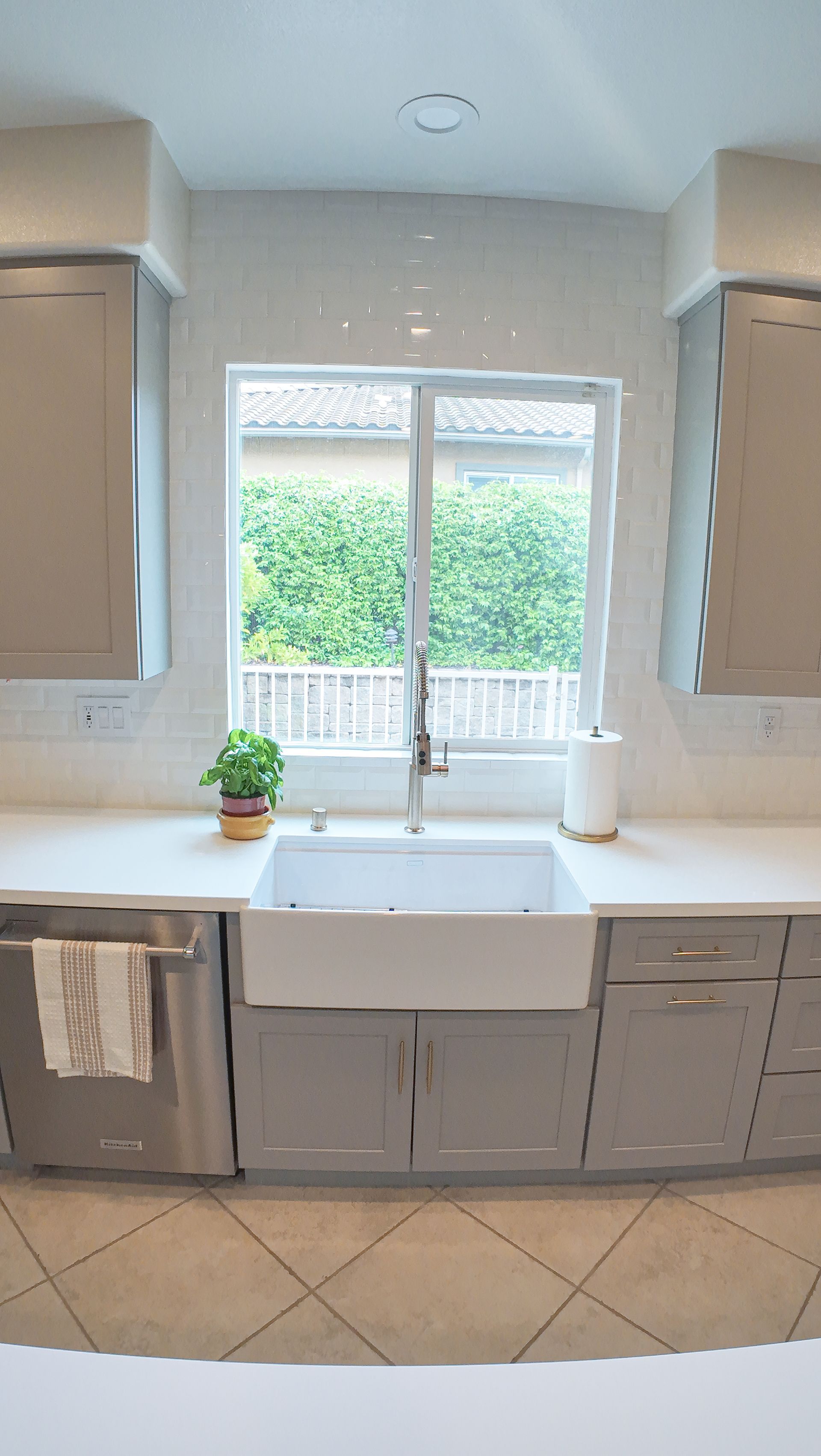 Kitchen with white farmhouse sink, grey cabinets, white backsplash, and a window overlooking greenery.