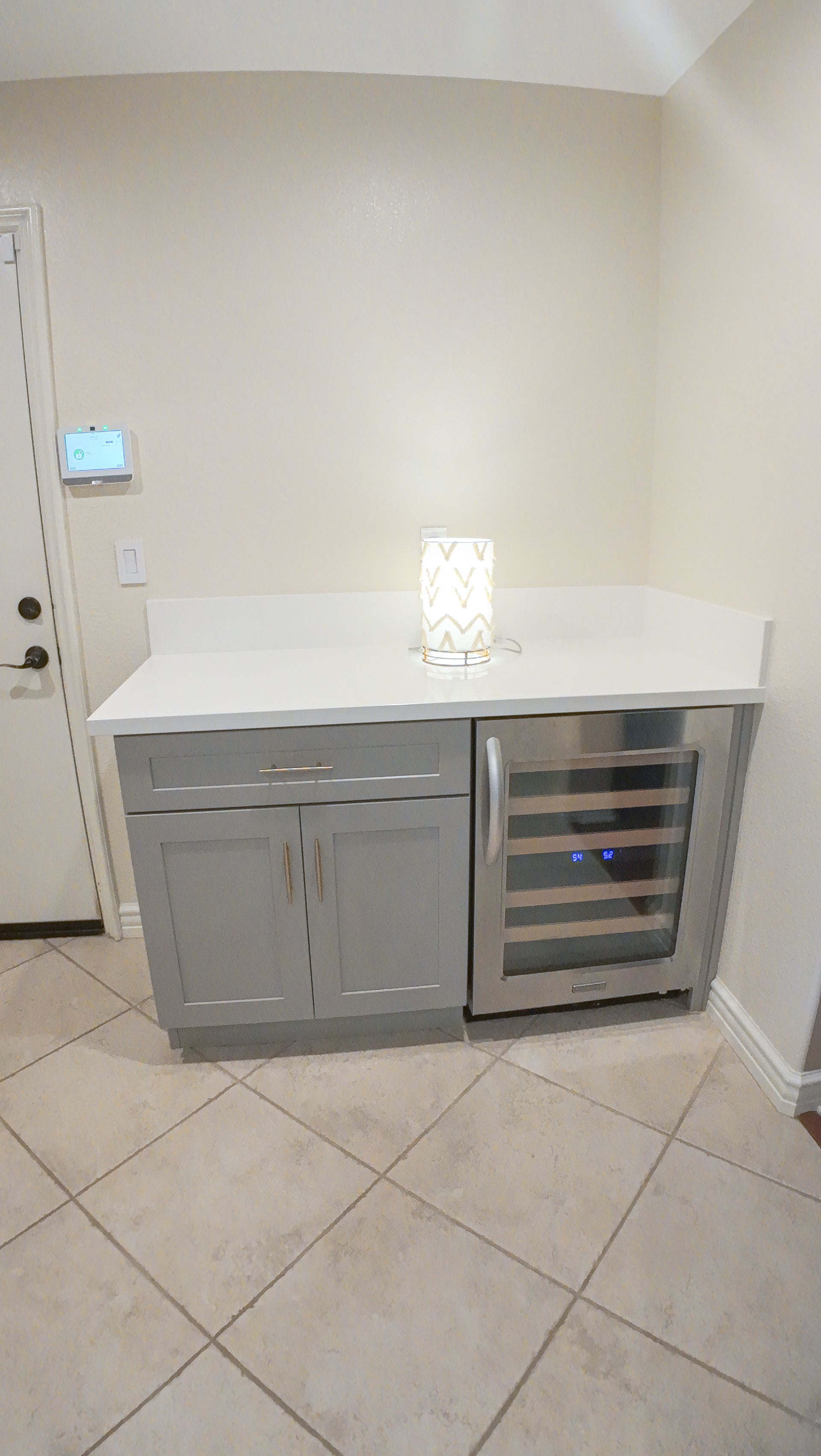 Gray cabinet with a built-in wine cooler and white countertop, with a lamp, against a beige wall.